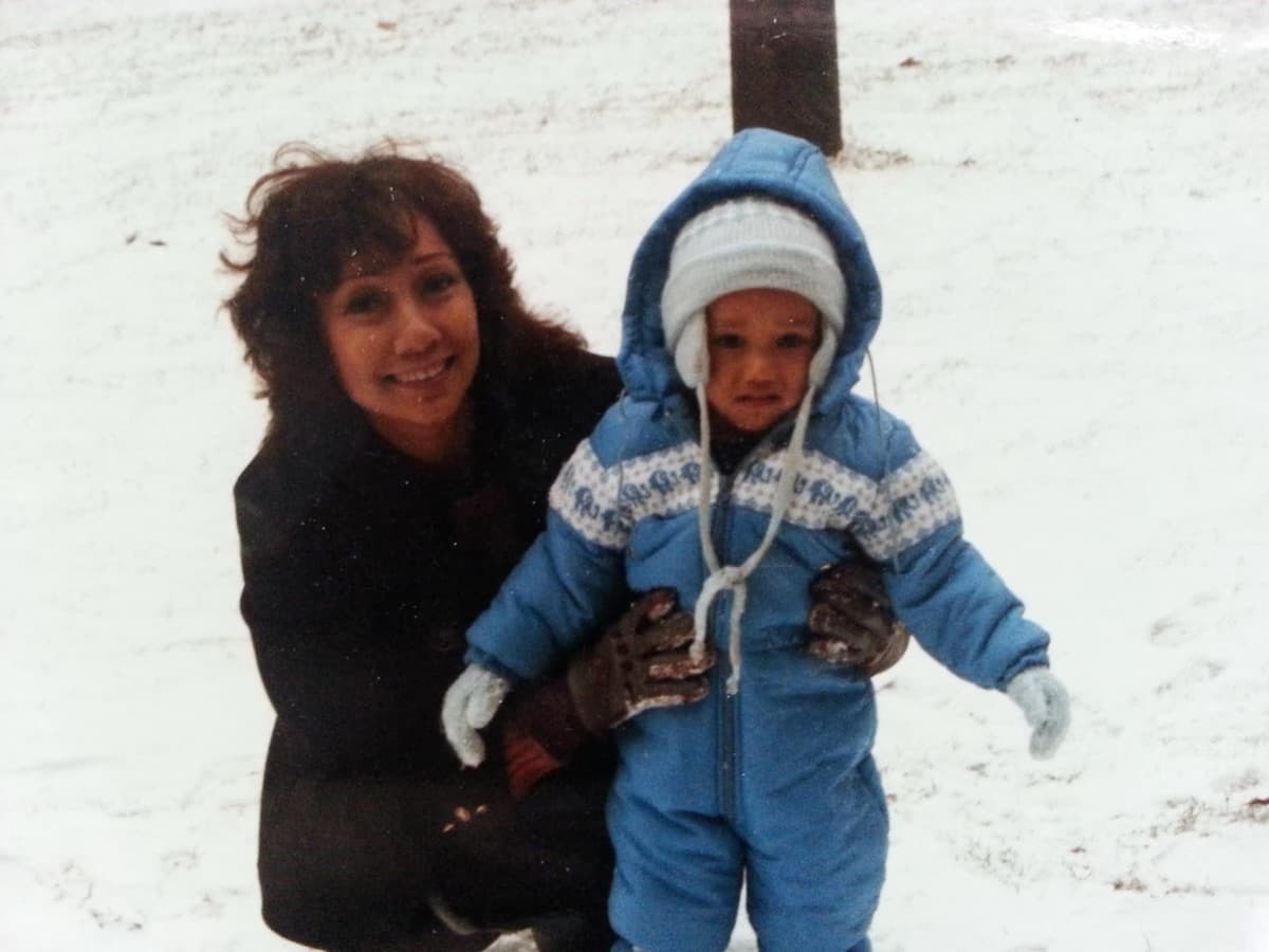 My mom crouched down beside me in the snow, holding onto me. I am small, in a blue snowsuit.
